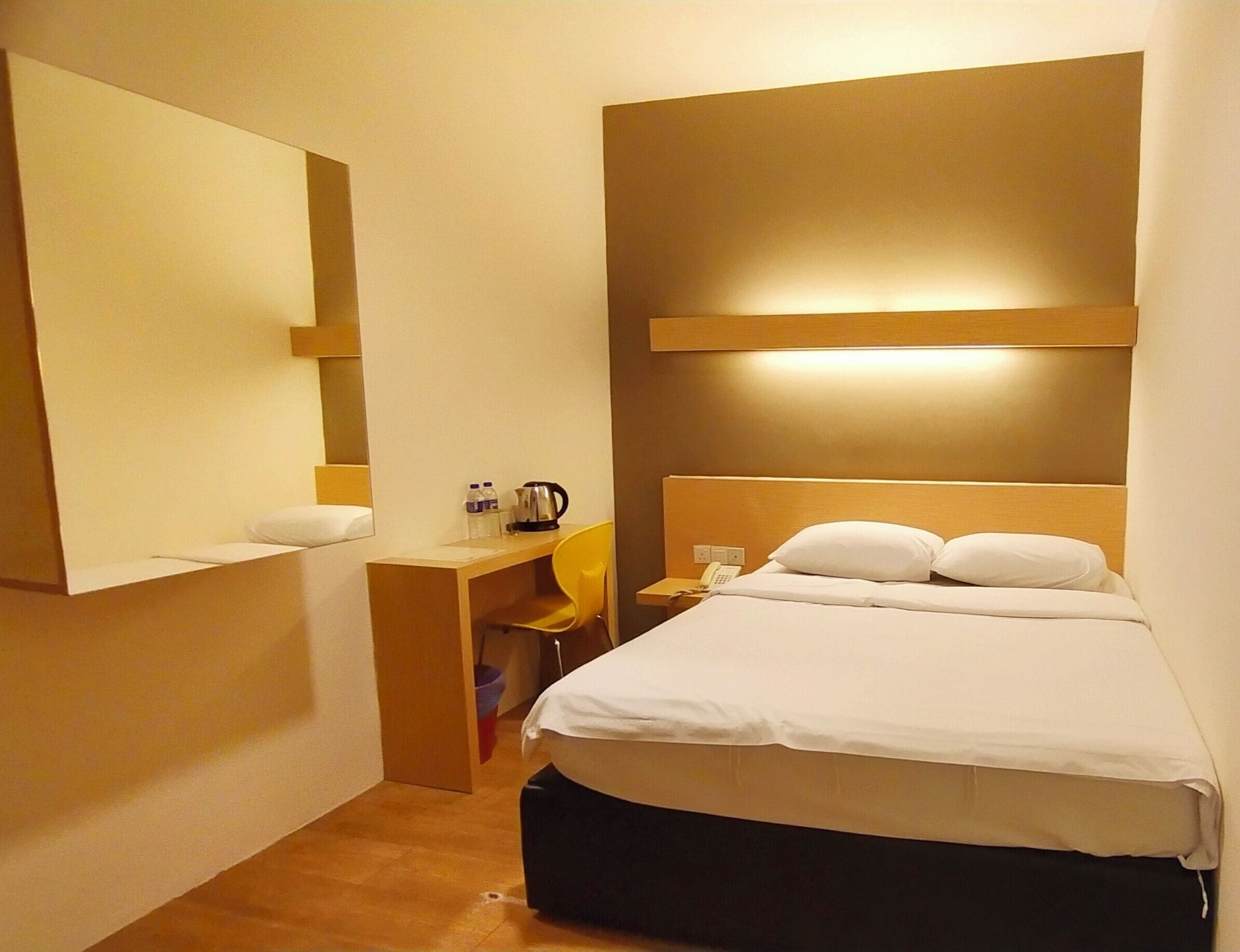 Une chambre d’hôtel équipée d’un lit, d’un bureau, d’une chaise, d’un miroir et d’une table de chevet avec un téléphone et une bouilloire.