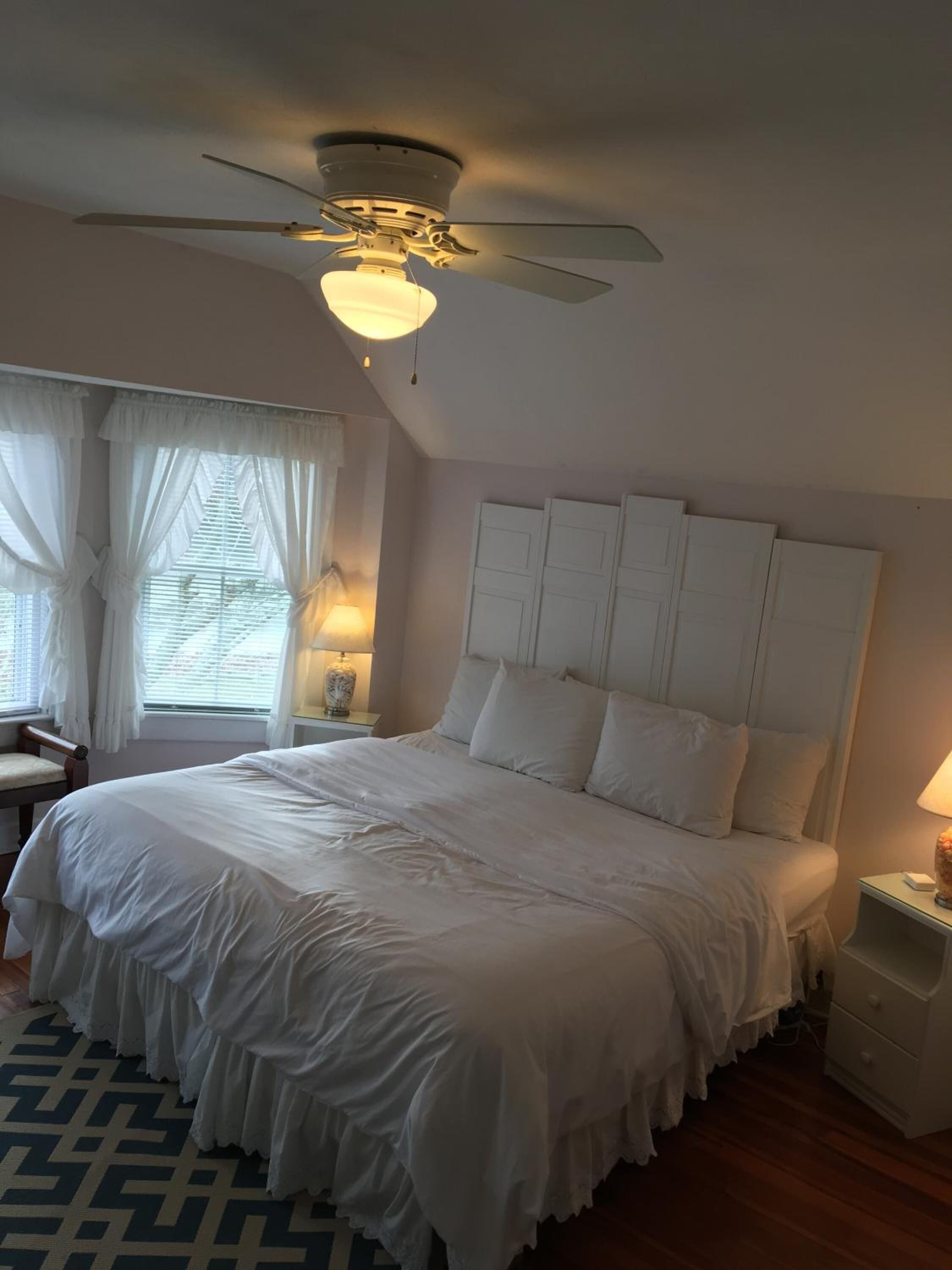 Une chambre à coucher avec un grand lit, un ventilateur de plafond, une table de chevet et une fenêtre avec des rideaux.