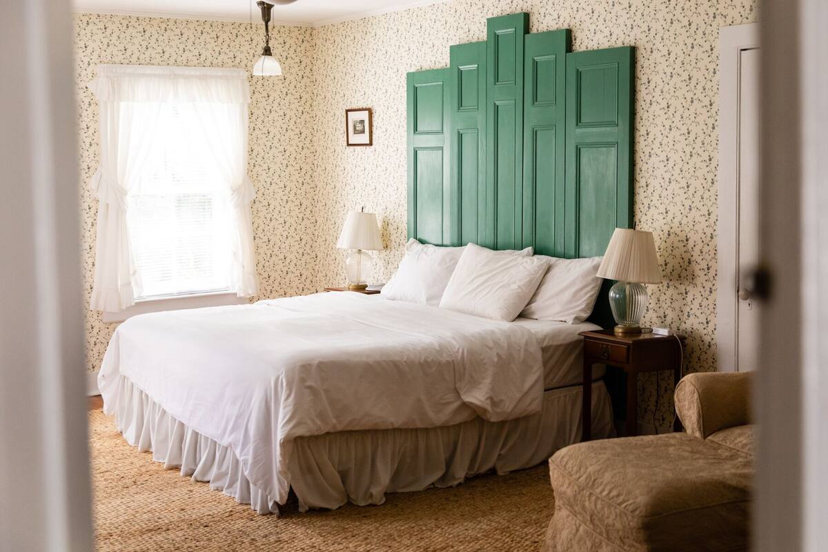 Un dormitorio con una cama grande, dos mesitas de noche con lámparas, un cabecero verde, papel tapiz floral y una ventana con cortinas.