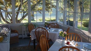 A conservatory with a dining table set for four, surrounded by wooden chairs, with a view of trees and a lawn. - House on Chase Creek (Shelter Island Heights)