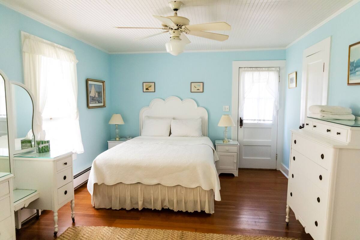 Un dormitorio con cama, dos mesitas de noche, cómoda y ventilador de techo, además de una puerta.