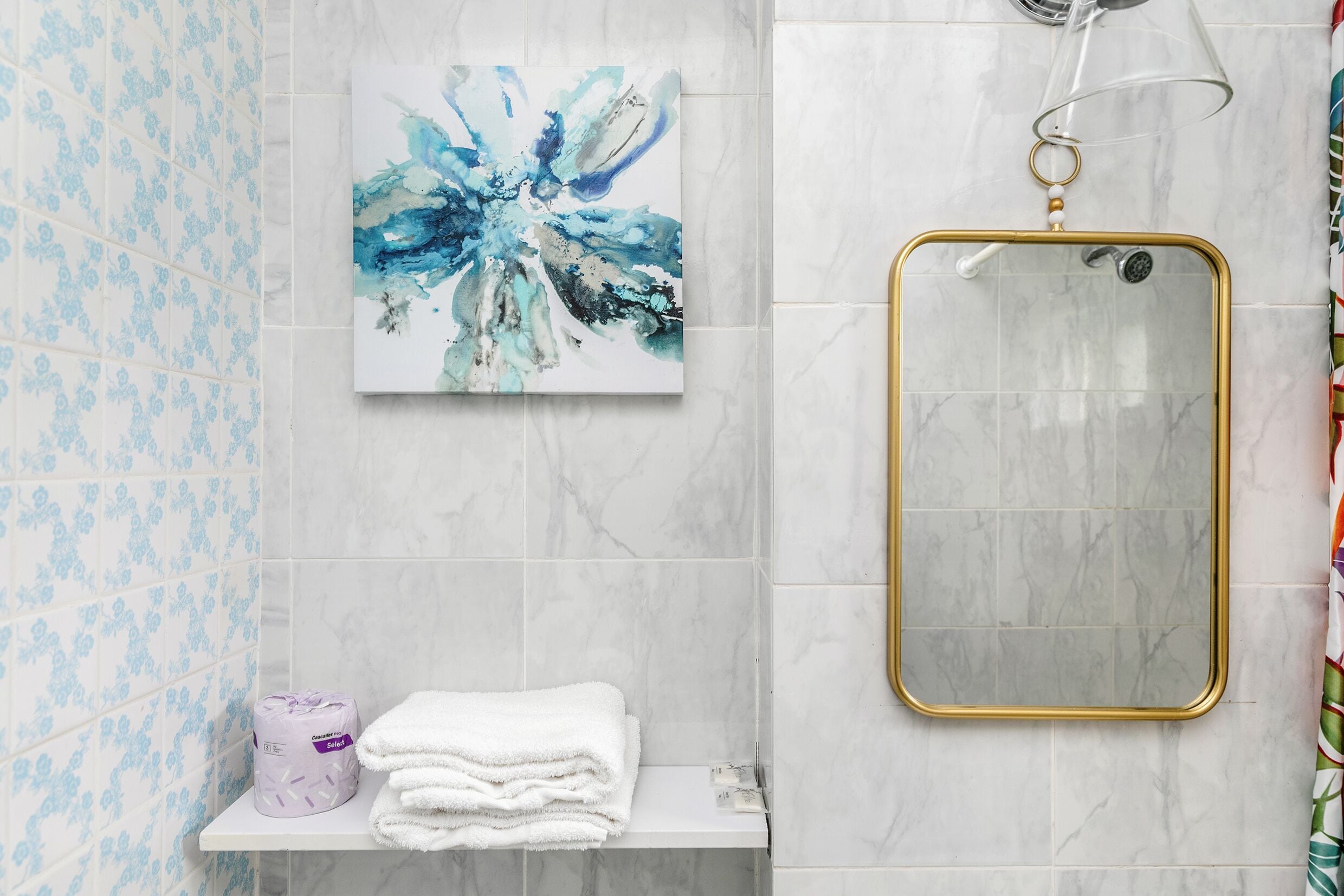 A bathroom with a marble wall, a blue floral-patterned tile backsplash, a white shelf with towels and toilet paper, and a gold-framed mirror.