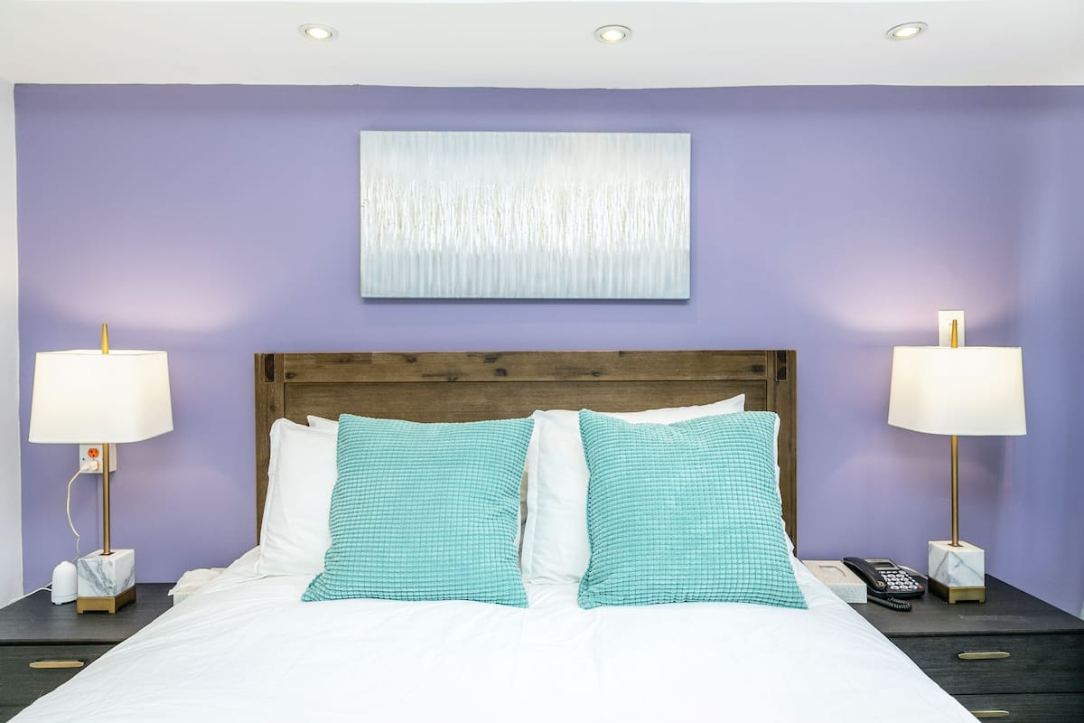 A hotel room with a large bed, two bedside lamps, a wooden headboard, a wall-mounted artwork, and a nightstand with a phone and a box.