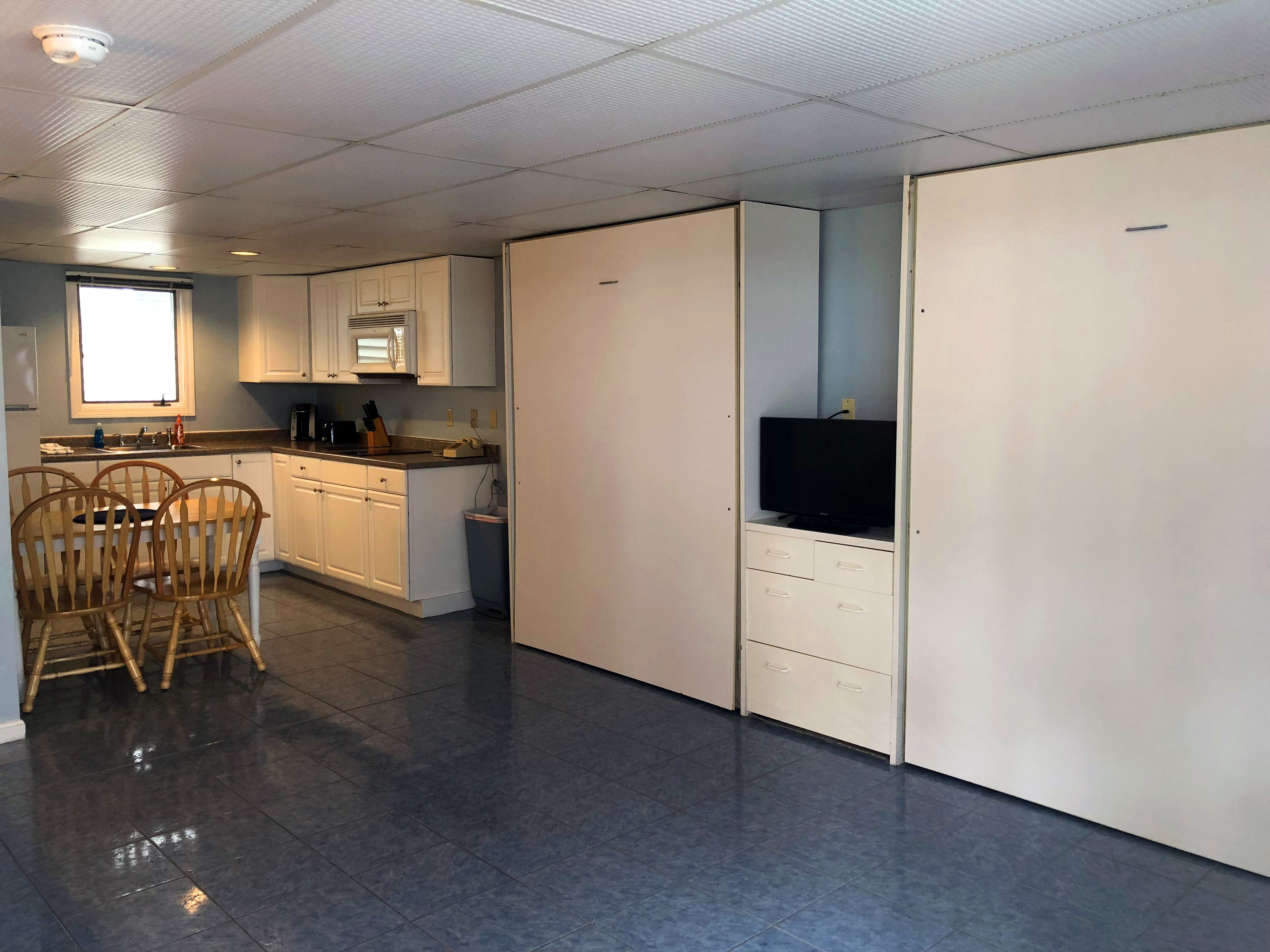 A modern kitchen with white cabinets, a dining area with wooden chairs, and a TV on a cabinet.