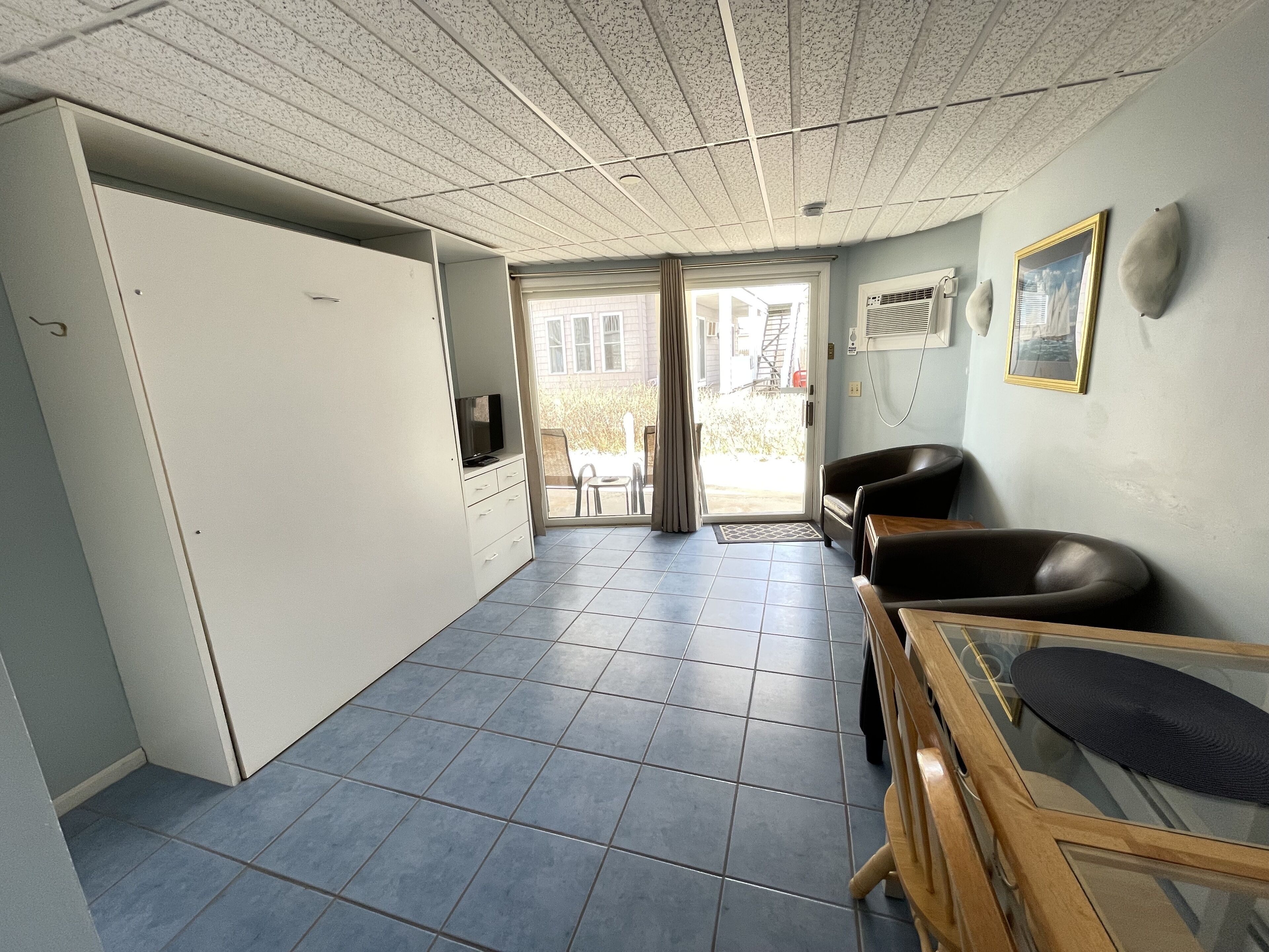 A room with a tiled floor, a television on a cabinet, a chair, a sofa, and a window with a view of the outside.