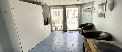A room with a tiled floor, a television on a cabinet, a chair, a sofa, and a window with a view of the outside.