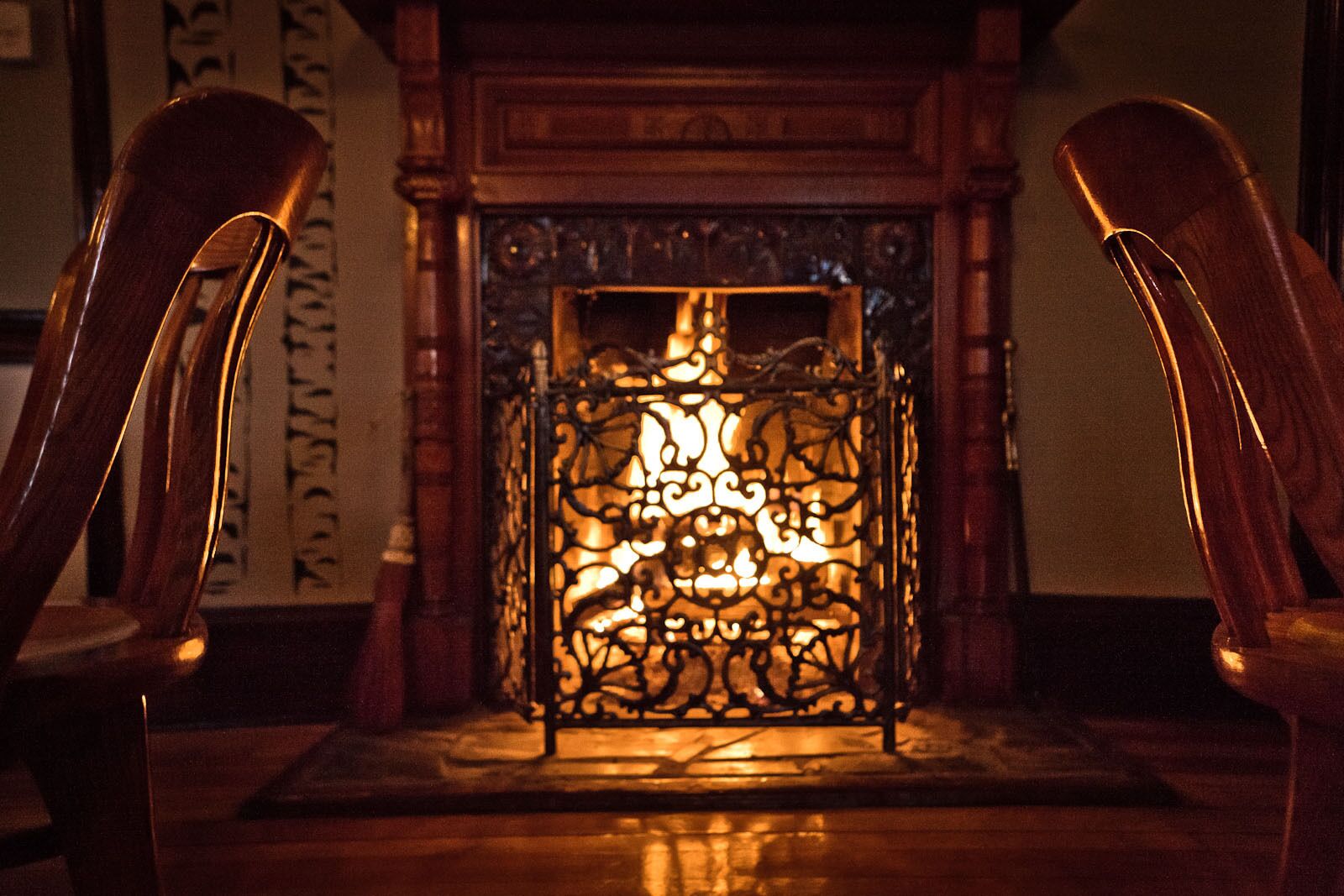 A fireplace with intricate metalwork, flanked by wooden chairs.