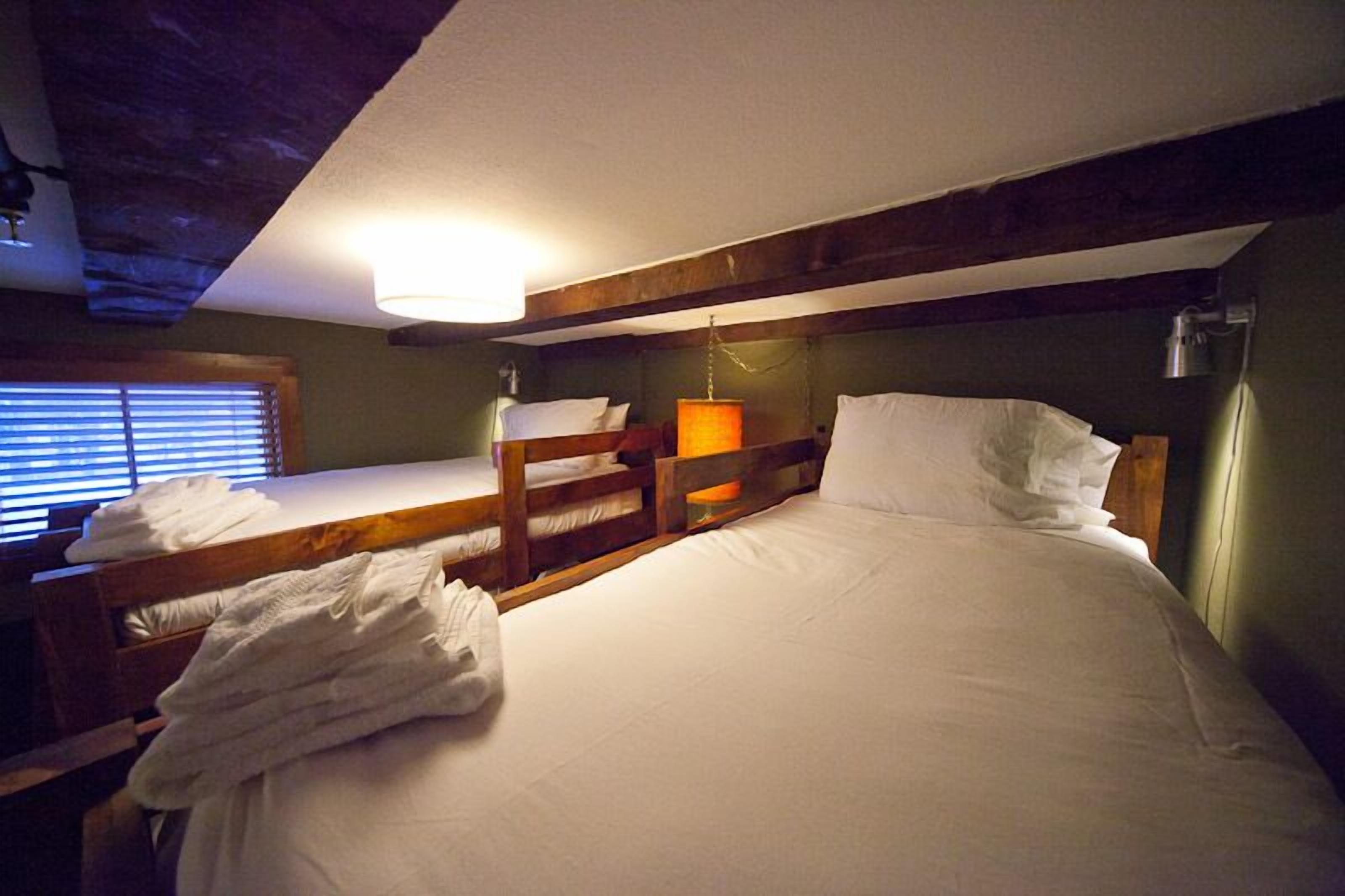 A room with bunk beds, a bedside table with a lamp, and a window with blinds.