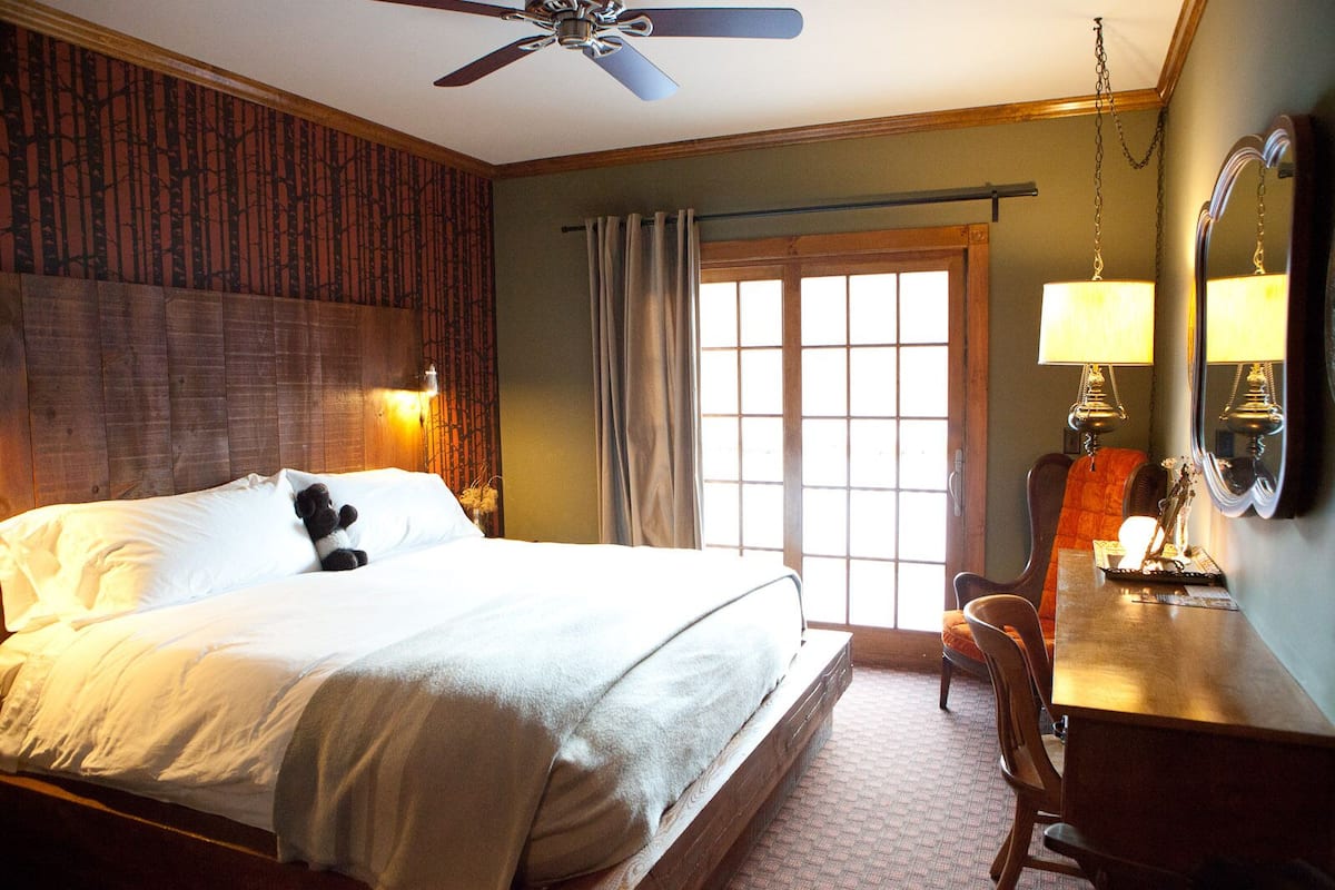 A hotel room with a bed, a desk, a chair, a ceiling fan, a window with curtains, and a wall-mounted lamp.