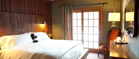A hotel room with a bed, a desk, a chair, a ceiling fan, a window with curtains, and a wall-mounted lamp.