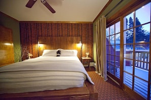 A large bed in a room with a wooden headboard, two bedside lamps, and a view of trees through large windows.
