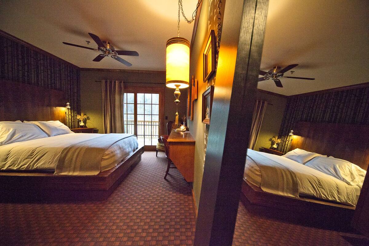A hotel room with two beds, a desk, a chair, a desk lamp, a ceiling fan, and a window.