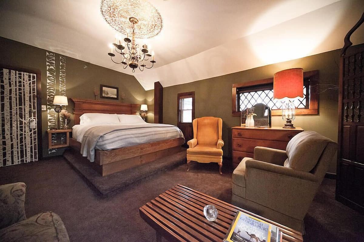 A bedroom with a large bed, two armchairs, a wooden coffee table, a chandelier, and a window with a red curtain.