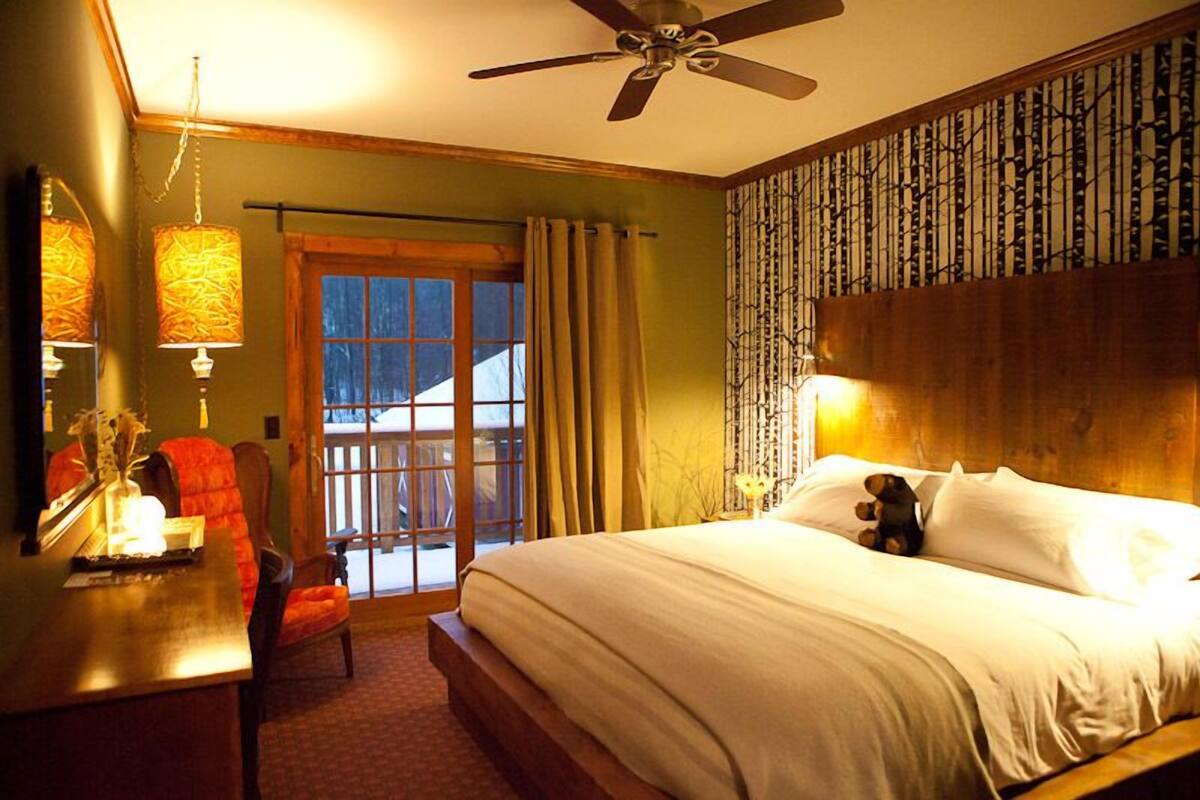 A bedroom with a bed, a desk, a chair, a ceiling fan, and a view of the outdoors through a window.