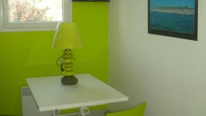 Una habitación pequeña con una mesa blanca, una silla verde, una lámpara verde, un televisor negro en la pared y una ventana con vista al mar.