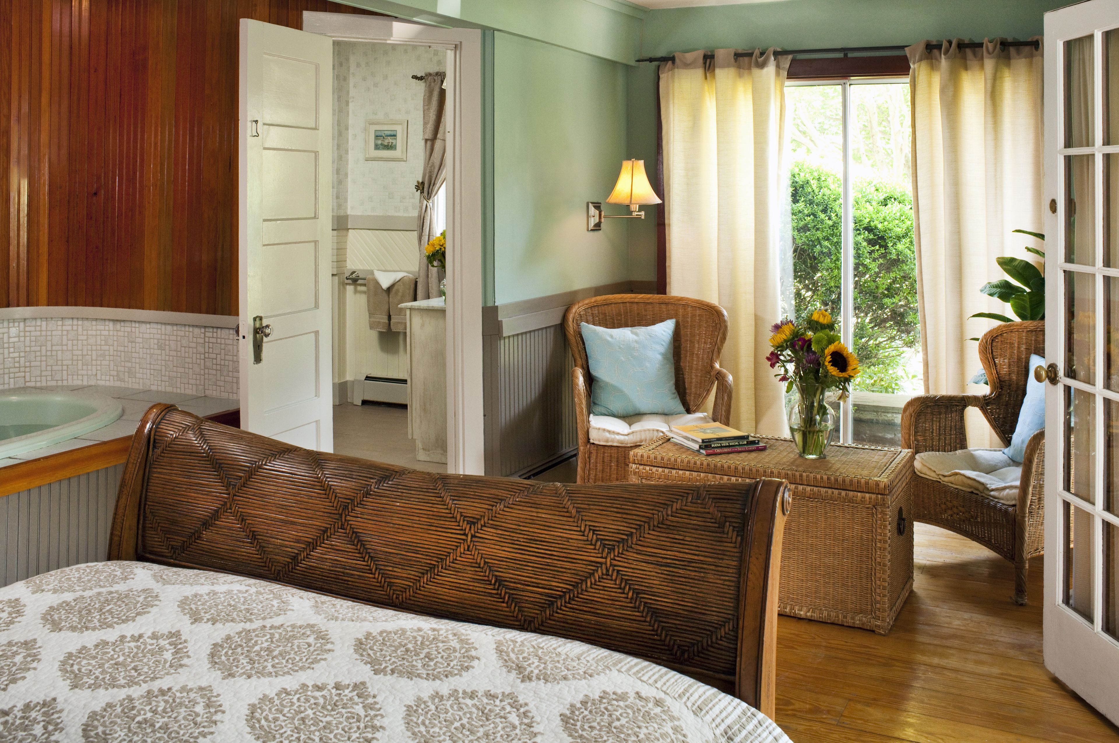 Un dormitorio con cama, una silla de mimbre, una mesita con un libro y flores, y vistas al exterior a través de una ventana.