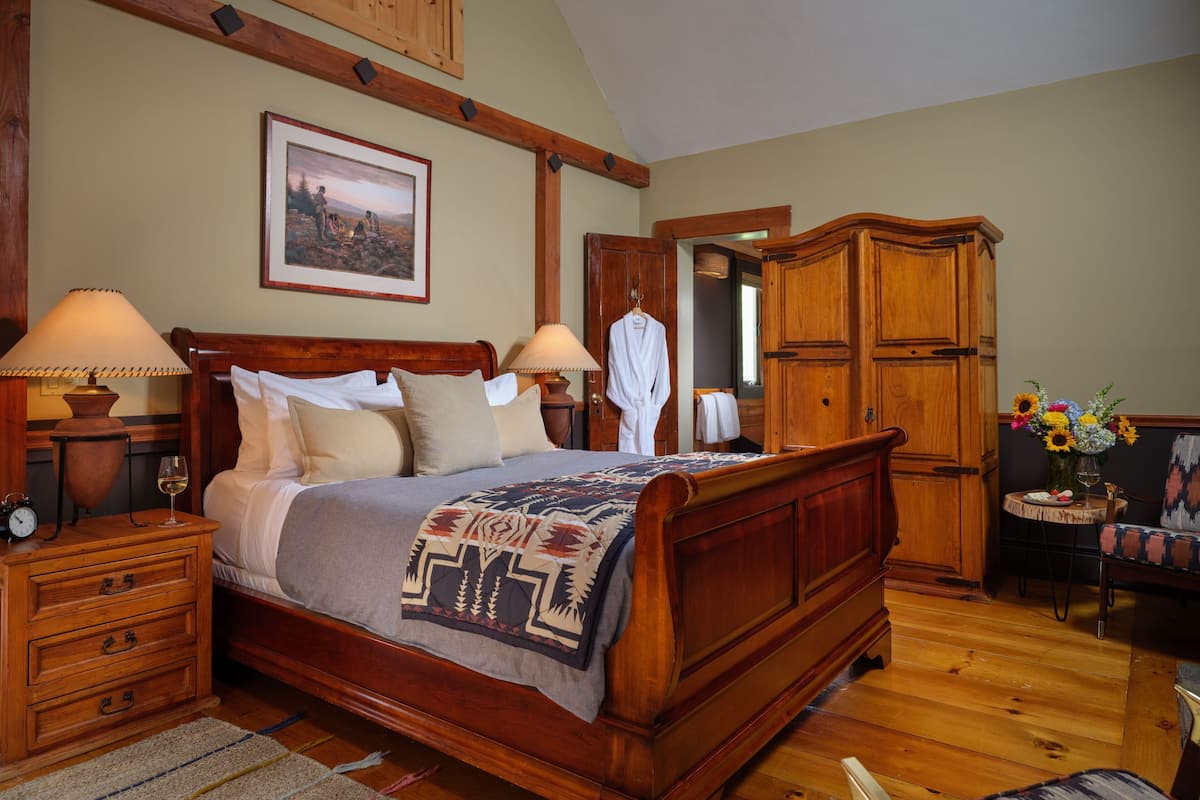A wooden four-poster bed with a patterned bedspread, a nightstand with a lamp, a dresser, and a small table with a vase of flowers.