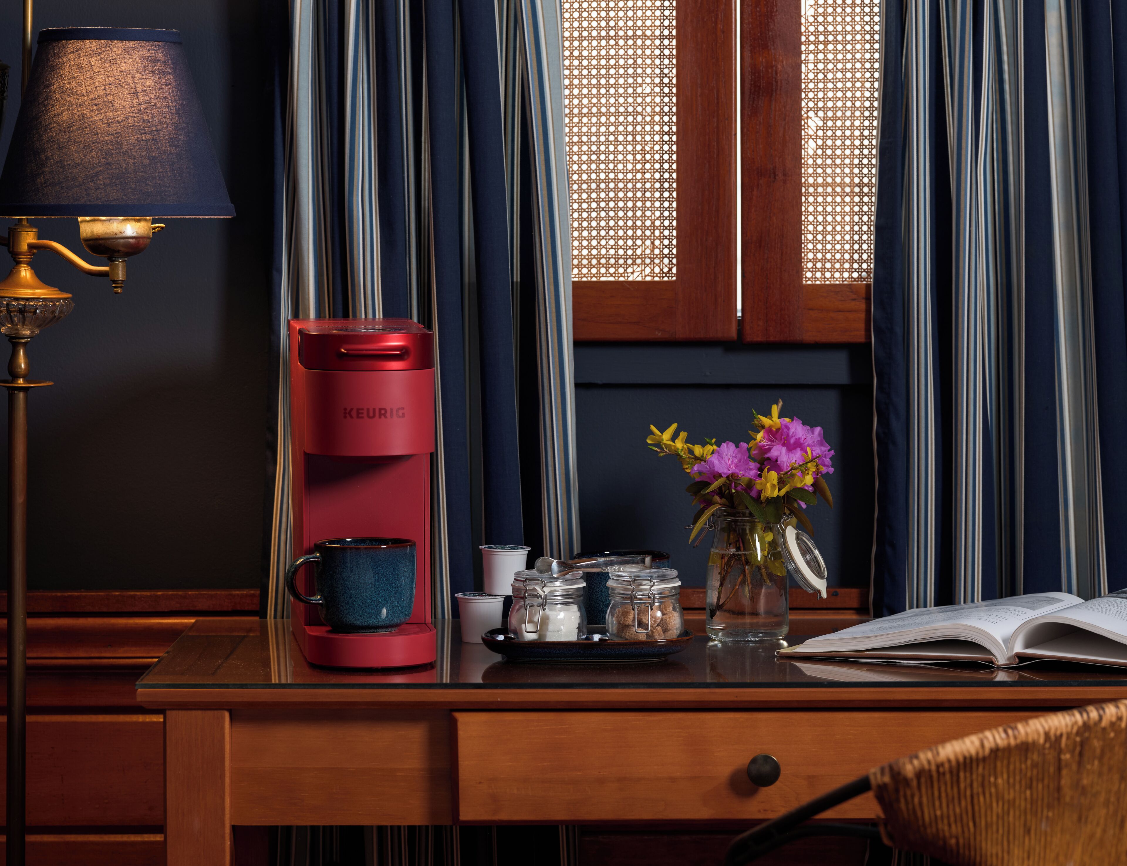 A desk with a red Keurig coffee maker, a blue mug, a glass jar with a metal lid, a vase with flowers, and an open book.