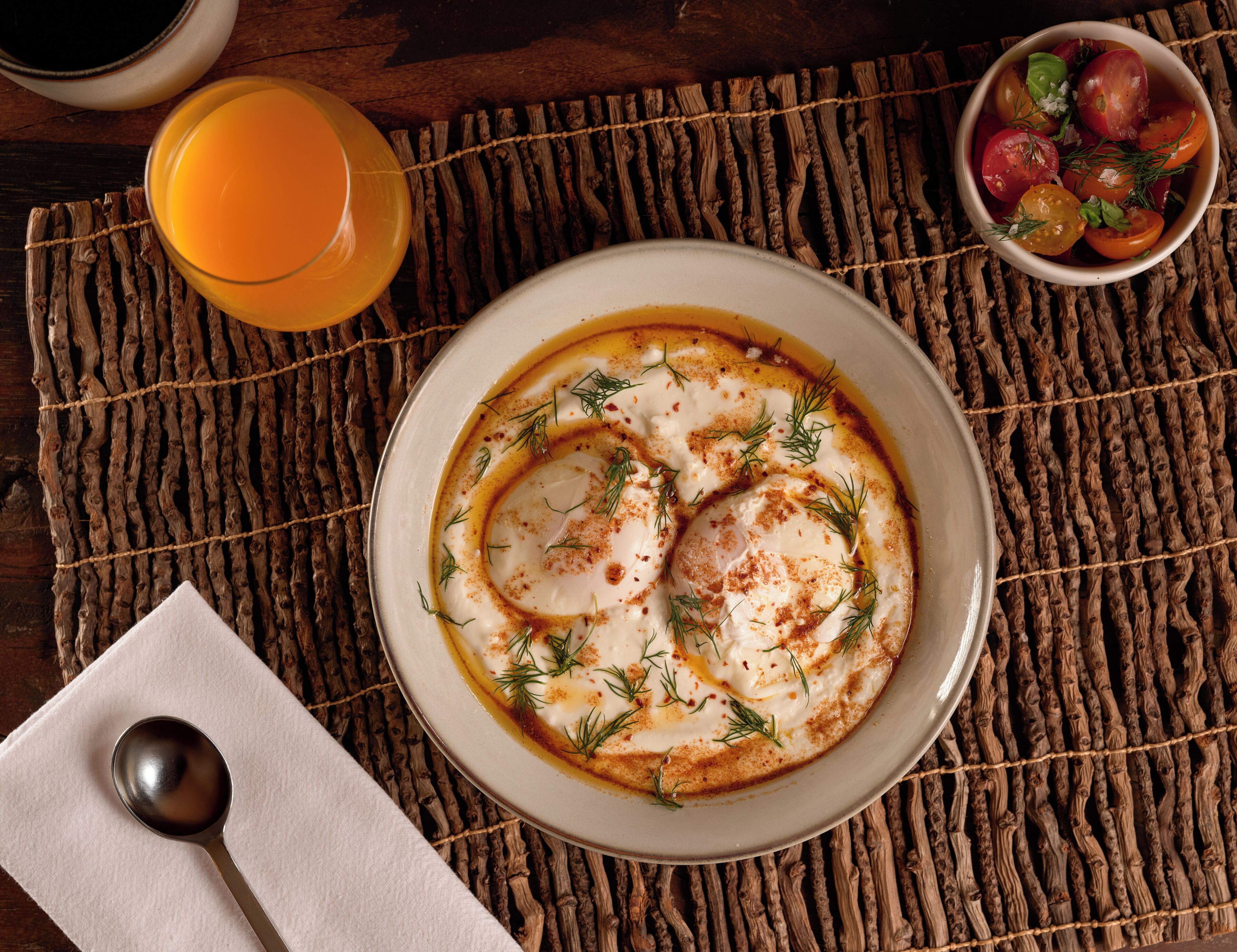 Uma tigela de comida sobre um apoio de palha trançada, uma colher, um copo de suco de laranja e uma pequena saladeira com frutas.