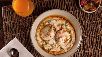 Uma tigela de comida sobre um apoio de palha trançada, uma colher, um copo de suco de laranja e uma pequena saladeira com frutas.
