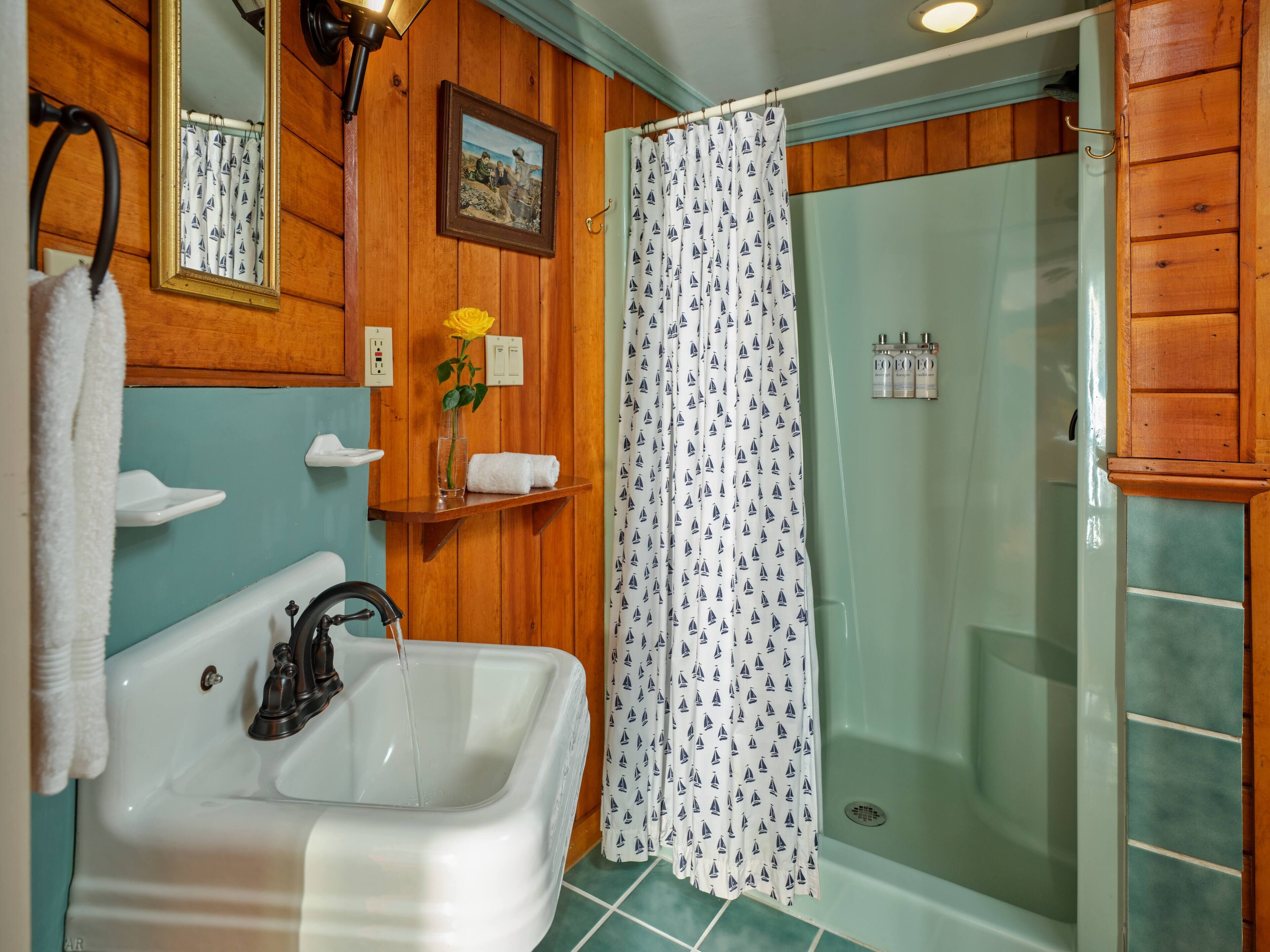 A bathroom with a white sink, a mirror, a shower with a sailboat curtain, and wooden paneling.