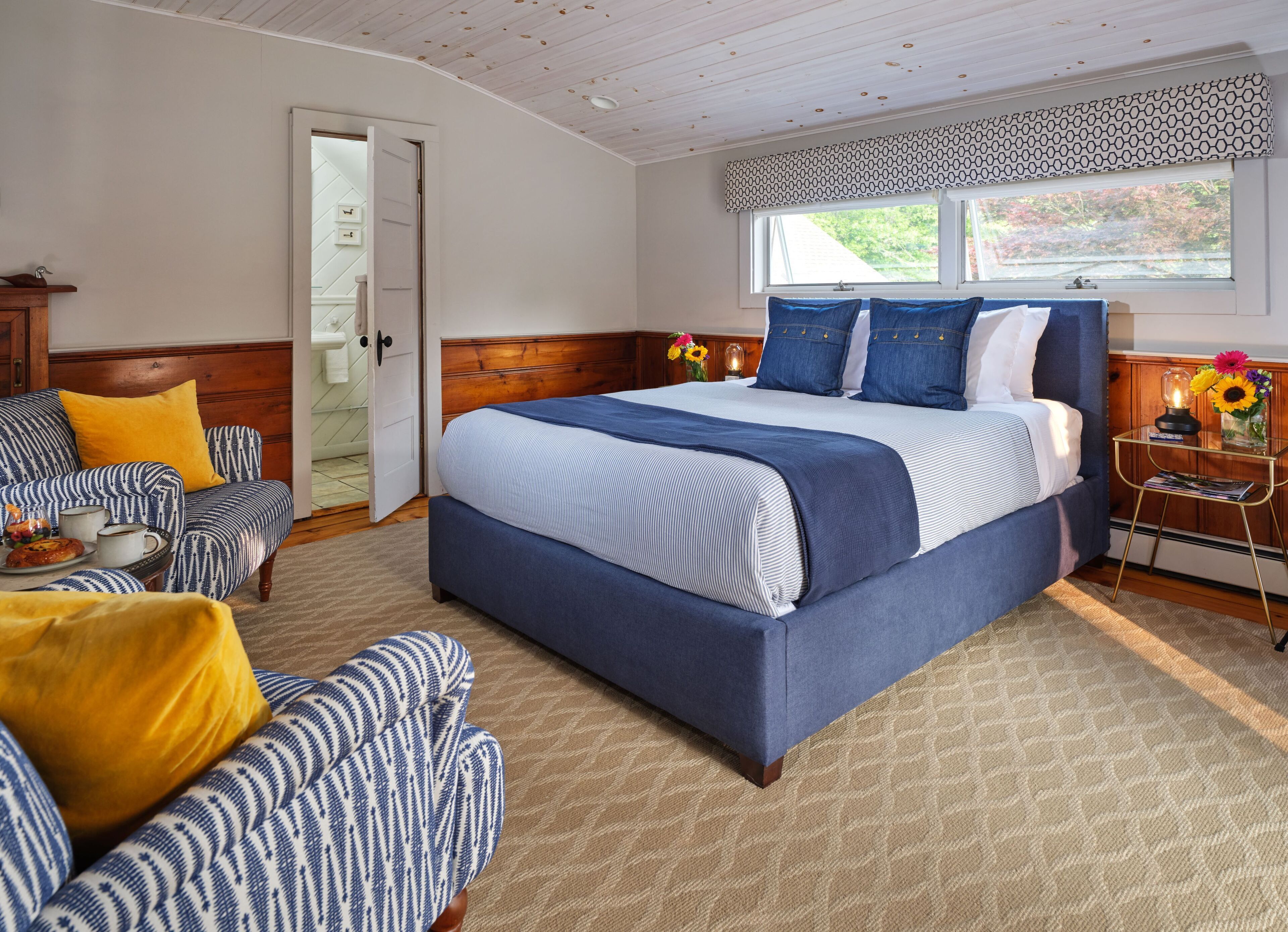 Une chambre avec un grand lit, une tête de lit en bois, une chaise avec un coussin jaune et une fenêtre agrémentée d’une lambrequin à motifs.