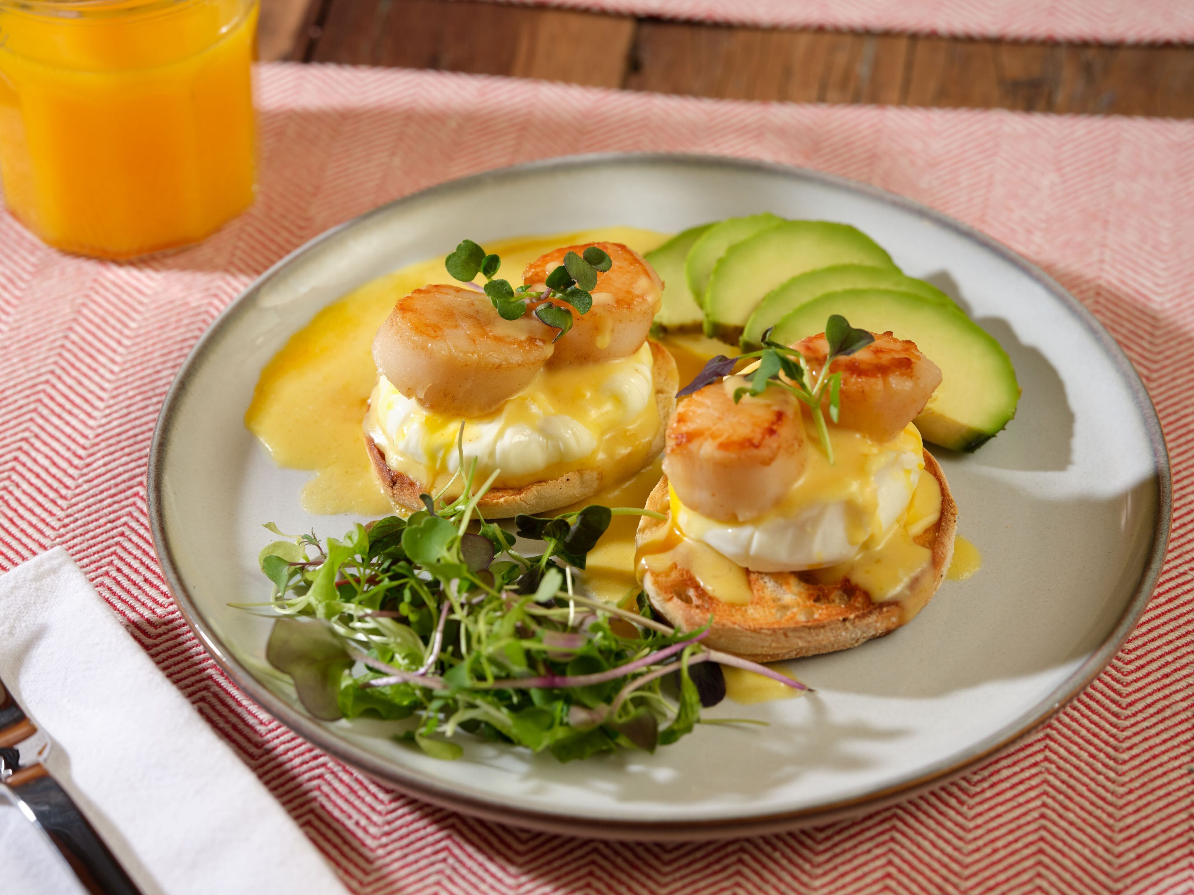 A plate with eggs benedict, avocado slices, and a glass of orange juice on a tablecloth.