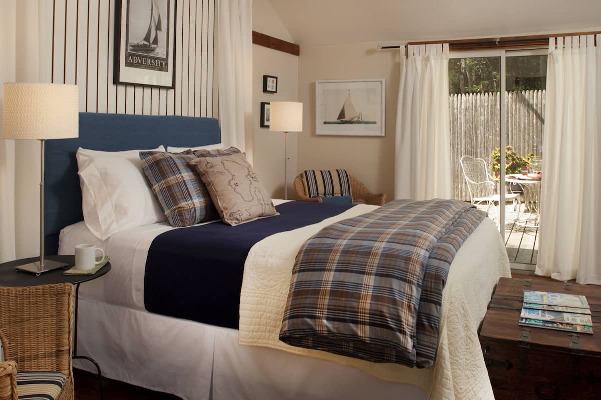 A neatly arranged hotel room with a large bed, a side table with a lamp, a wicker chair, and a view of the outdoors through a sliding door.