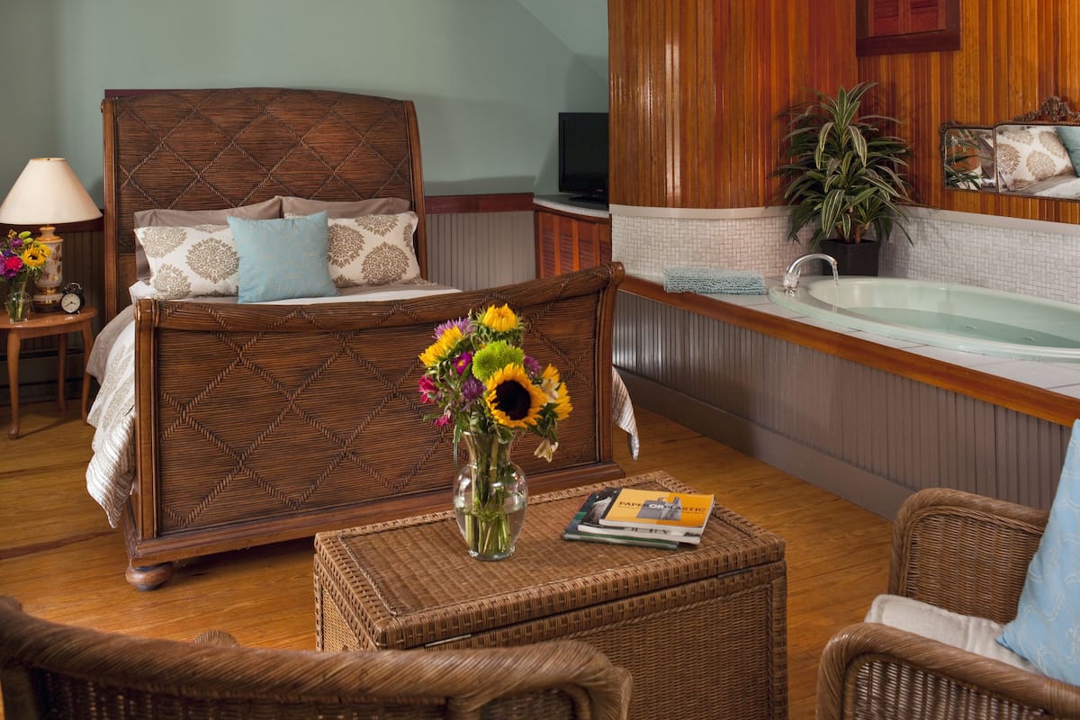 A bedroom with a wooden bed, a sitting area with wicker furniture, a small table with a vase of flowers, and a bathroom with a bathtub.