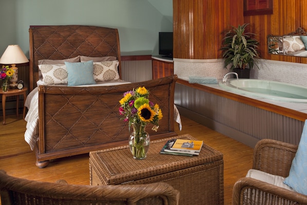 A bedroom with a wooden bed, a sitting area with wicker furniture, a small table with a vase of flowers, and a bathroom with a bathtub. - Abbey's Lantern Hill Inn (Ledyard)