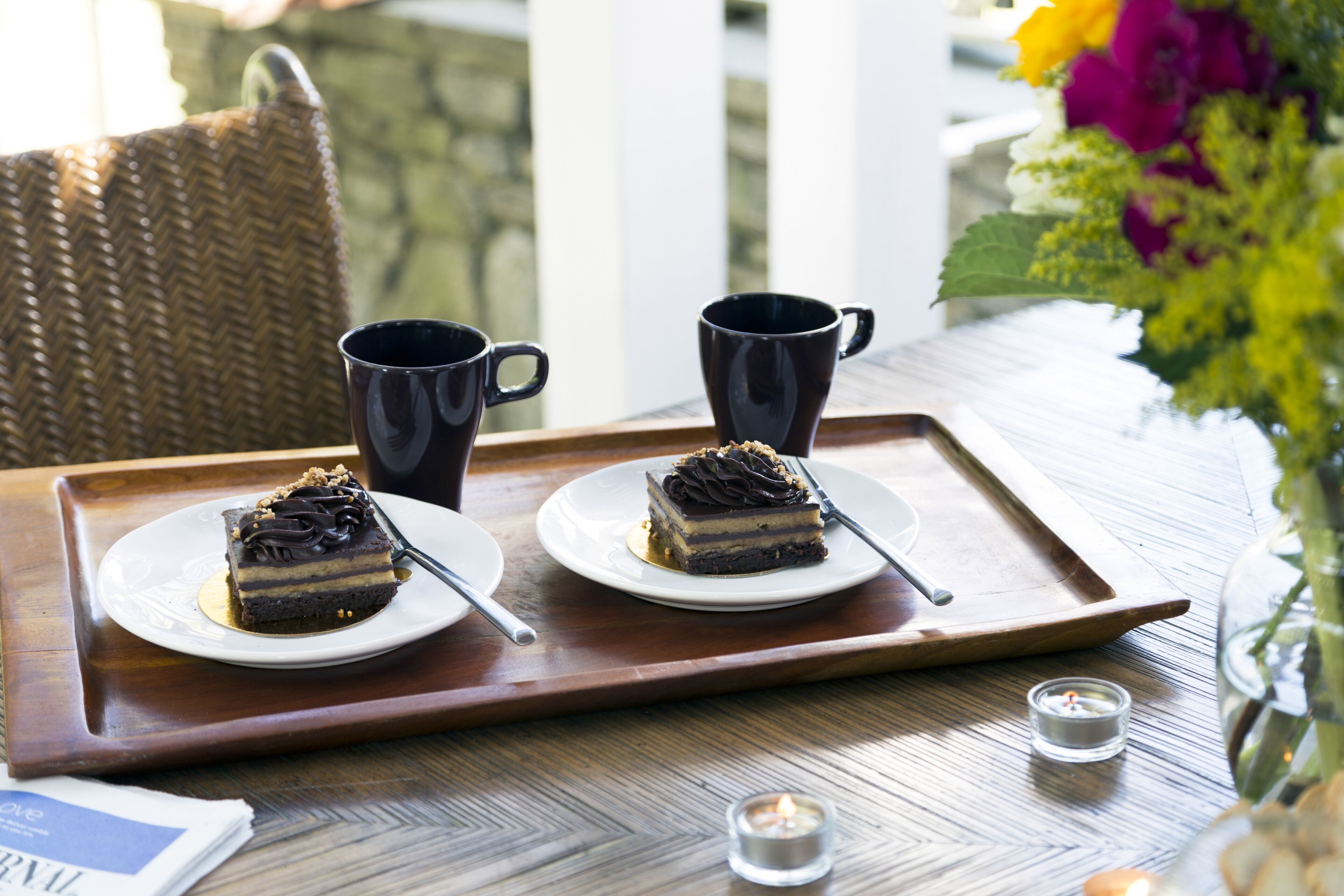 A tray with two plates of cake, two black mugs, and a candle on a wooden table.