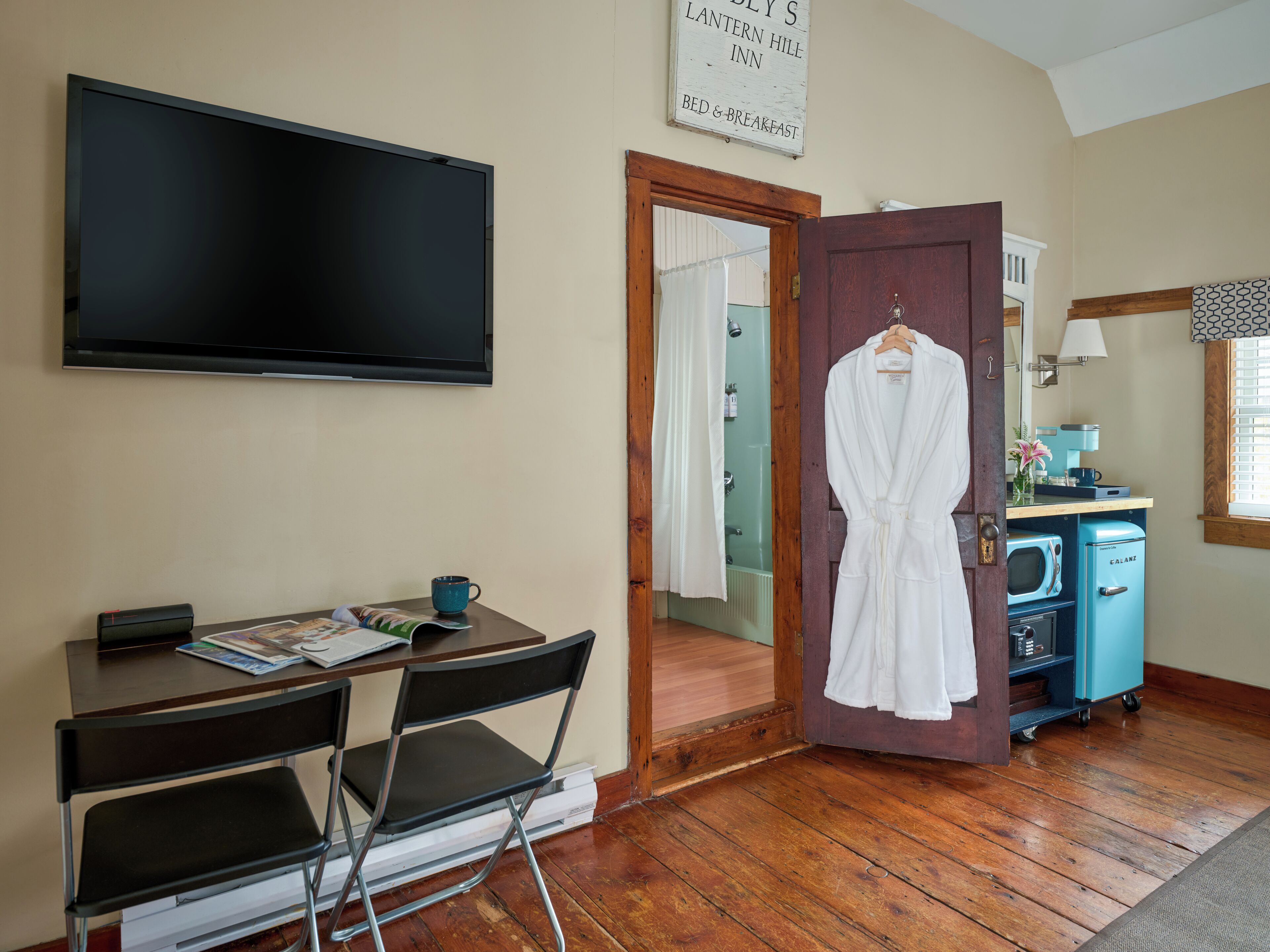 Ein Hotelzimmer mit Flachbildfernseher, Schreibtisch mit Stühlen, Bademantel an der Tür und einer Küchenzeile mit blauem Kühlschrank.