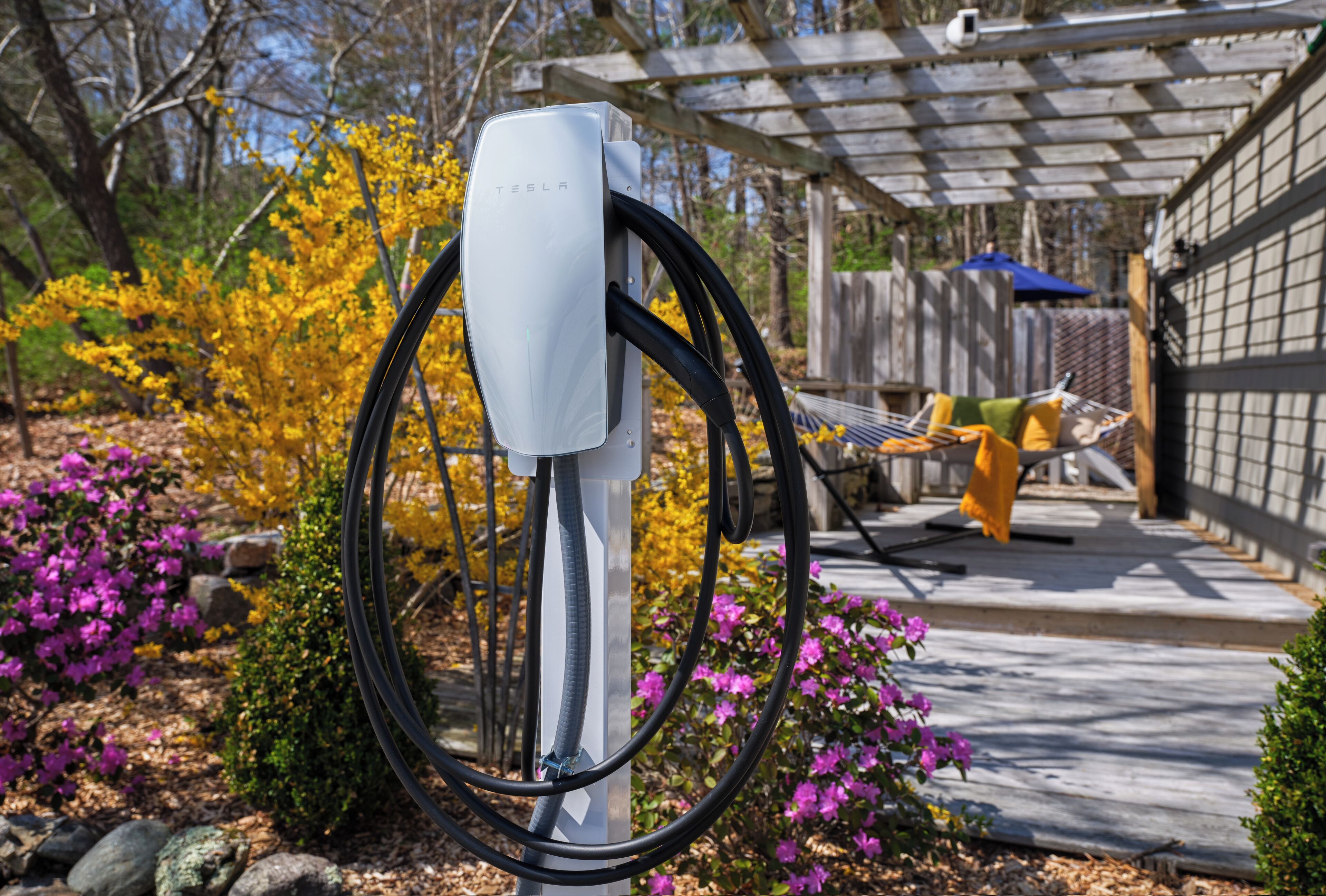 Estação de carregamento Tesla em um jardim com pérgola, área de estar externa e plantas floridas.