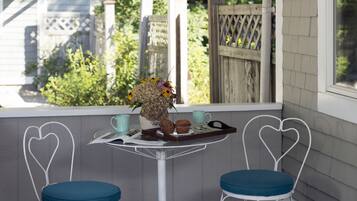 Um pequeno pátio com duas cadeiras em formato de coração, uma mesa com uma bandeja de muffins e um vaso de flores, e vista para uma cerca de madeira e vegetação.