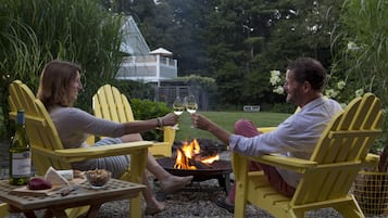Two people sitting on yellow Adirondack chairs at a campfire, with a bottle of wine and cheese on a table in front of them.