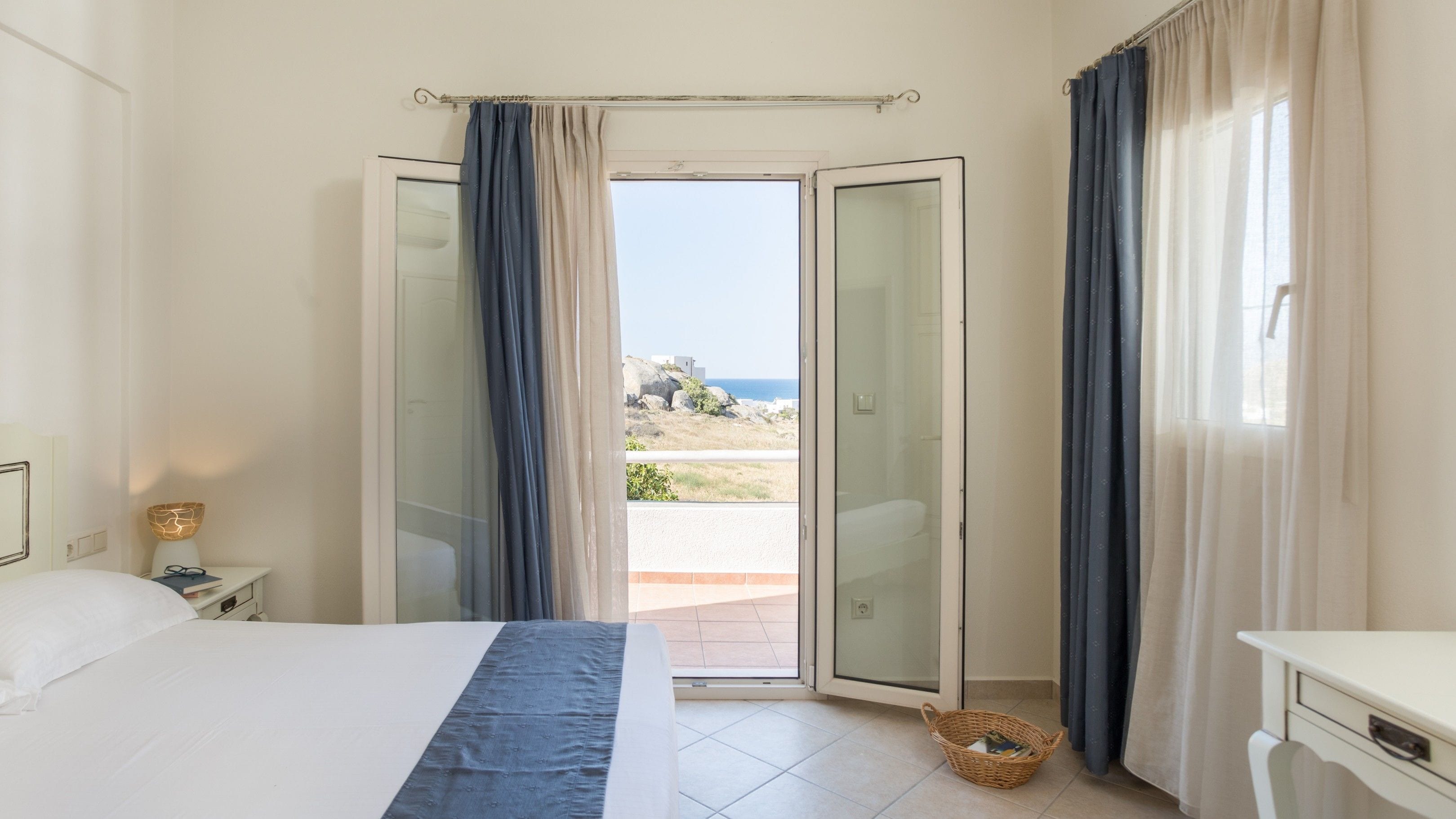 Ein Schlafzimmer mit einem Bett, einem Nachttisch, einer Lampe und Blick auf das Meer durch eine Glastür.
