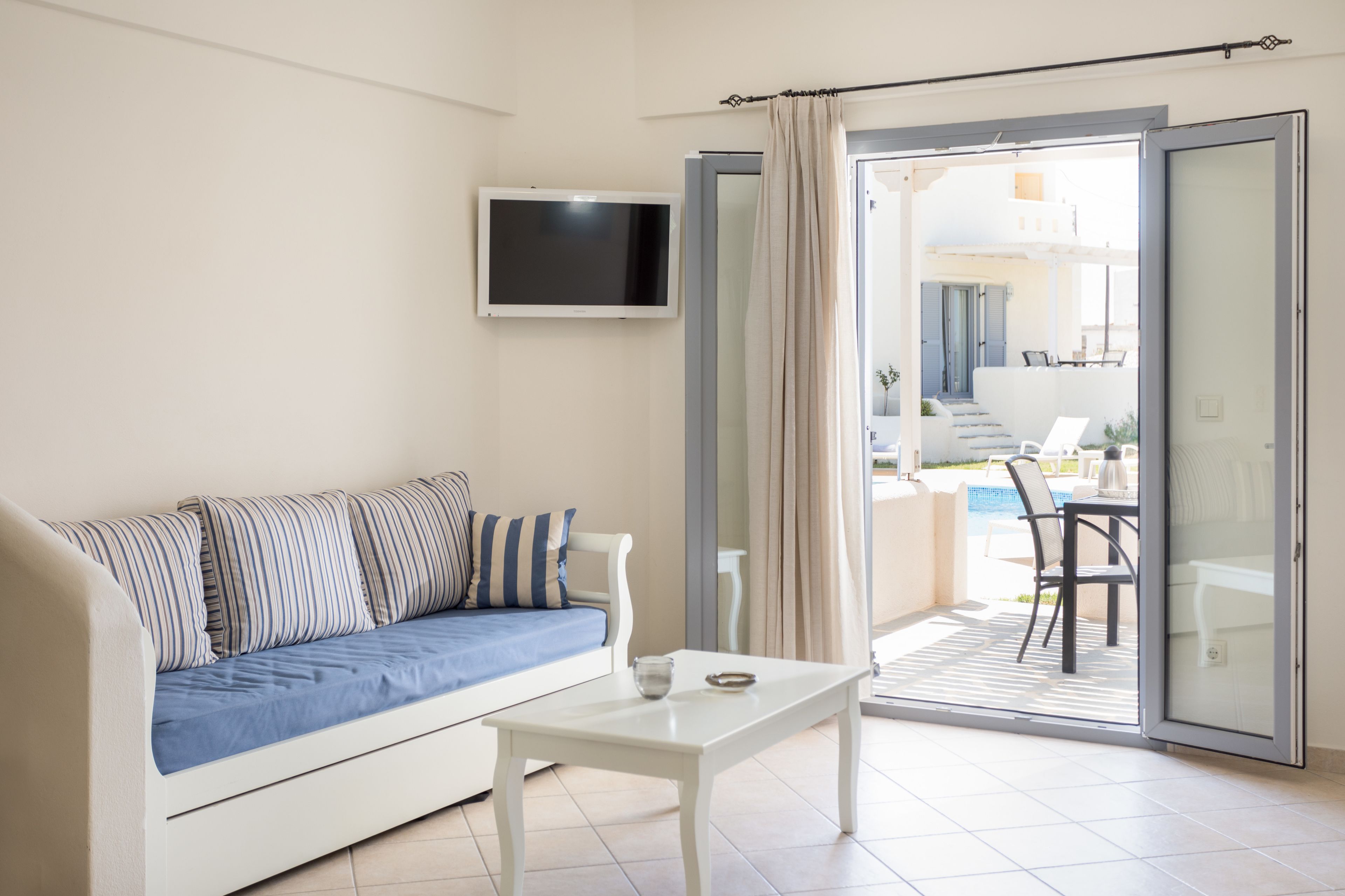 Sala de estar com sofá, mesa de centro e televisão. Há uma porta de vidro deslizante que dá para uma área externa com piscina e cadeiras.