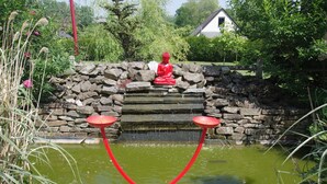 A red Buddha statue on a stone platform with a pond and two red stools in front. - Le Domaine de la Belle Normandie (Condé-en-Normandie)