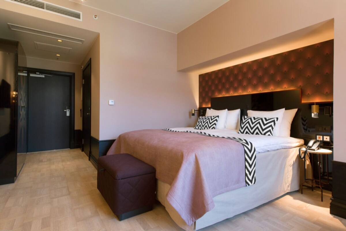 Une chambre d’hôtel moderne avec un grand lit, une table de chevet avec une lampe et un petit tabouret.