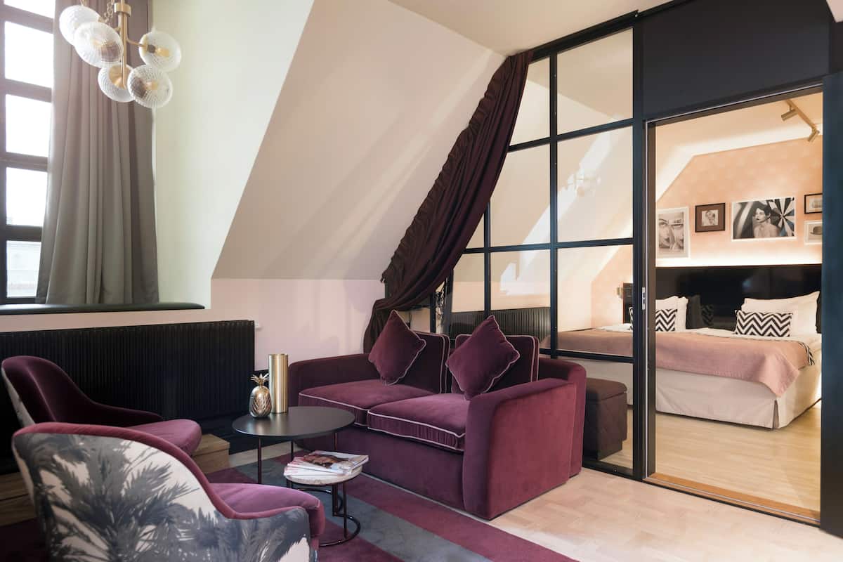 Un salon moderne avec un canapé, des fauteuils et une table basse. Une porte-fenêtre coulissante donne accès à une chambre à coucher équipée d’un lit et d’une table de chevet.