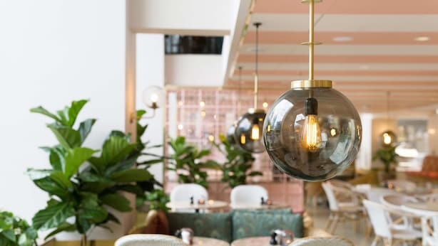 Un comedor moderno con mesas redondas y sillas de mimbre, con lámparas esféricas colgantes y plantas verdes.