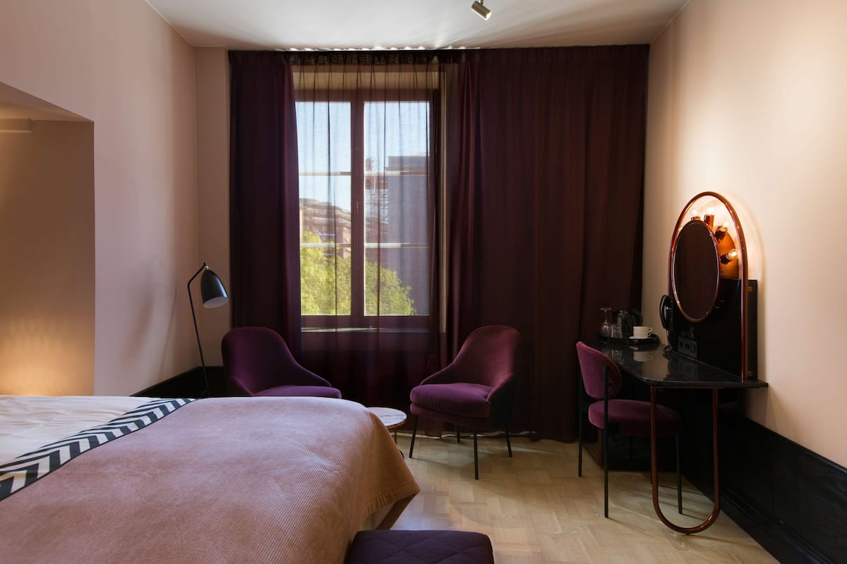 Une chambre d’hôtel avec un lit, deux chaises, un bureau avec un miroir et une fenêtre avec des rideaux.