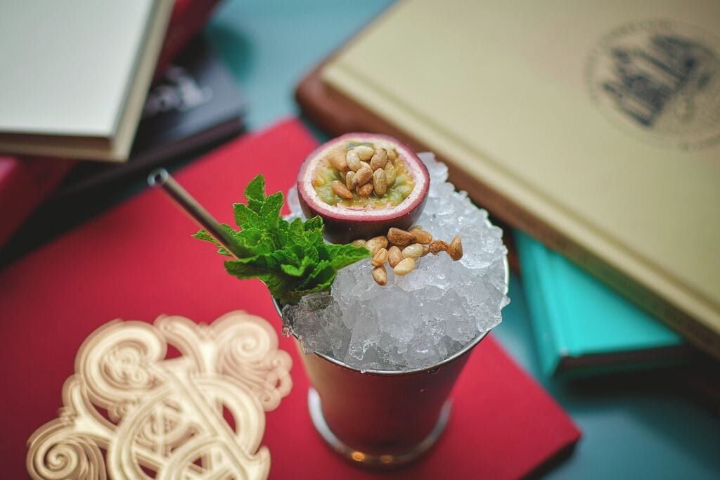 A cocktail with ice and a passion fruit slice garnished with nuts, placed on a red surface with books and a decorative item nearby.