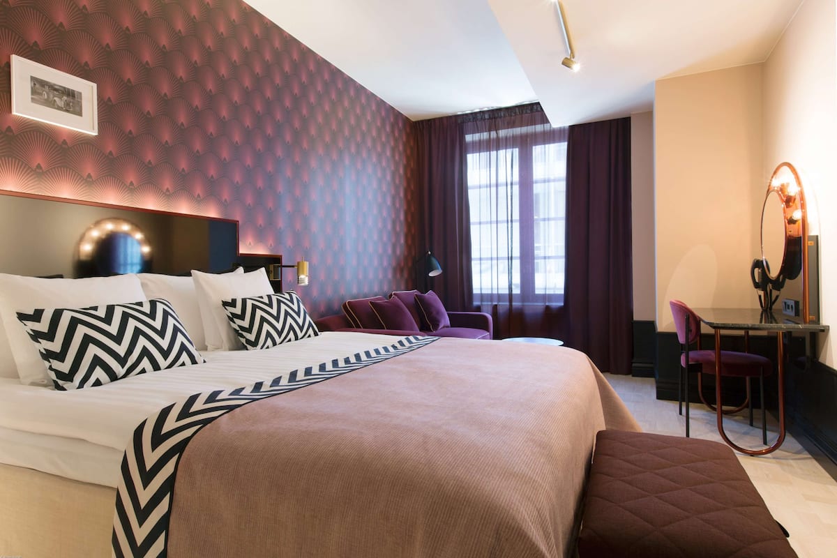 Une chambre d’hôtel avec un grand lit, un bureau avec une chaise, un miroir et une fenêtre avec des rideaux.