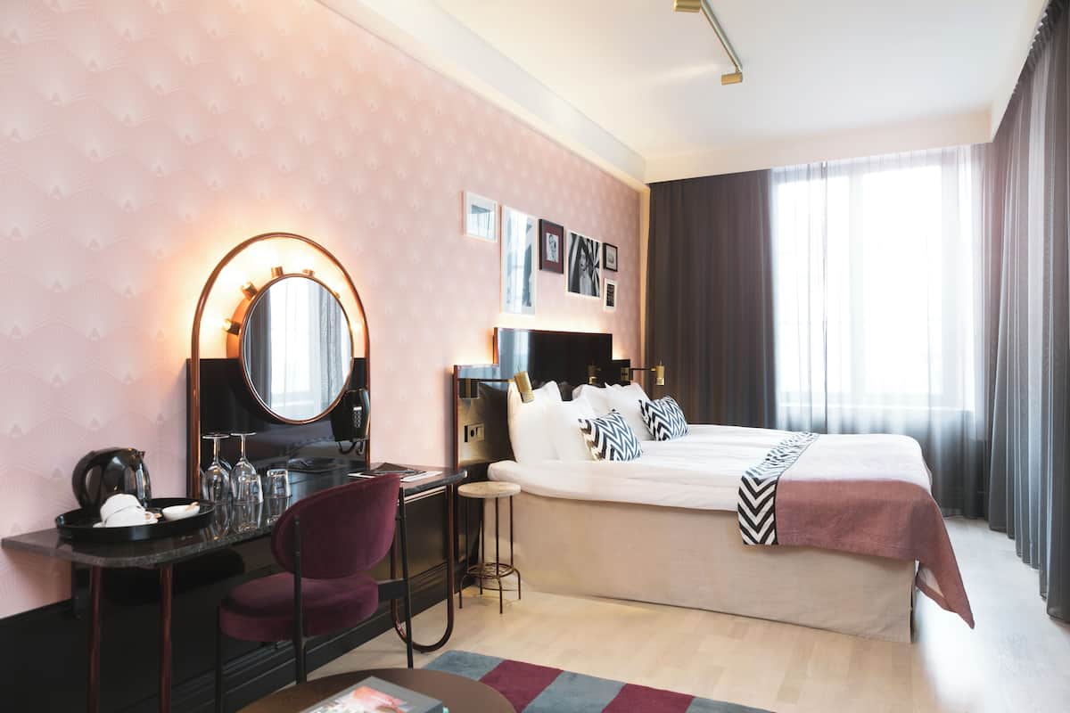 Une chambre d’hôtel avec un lit, un bureau et une chaise, un miroir rond et un petit tabouret.