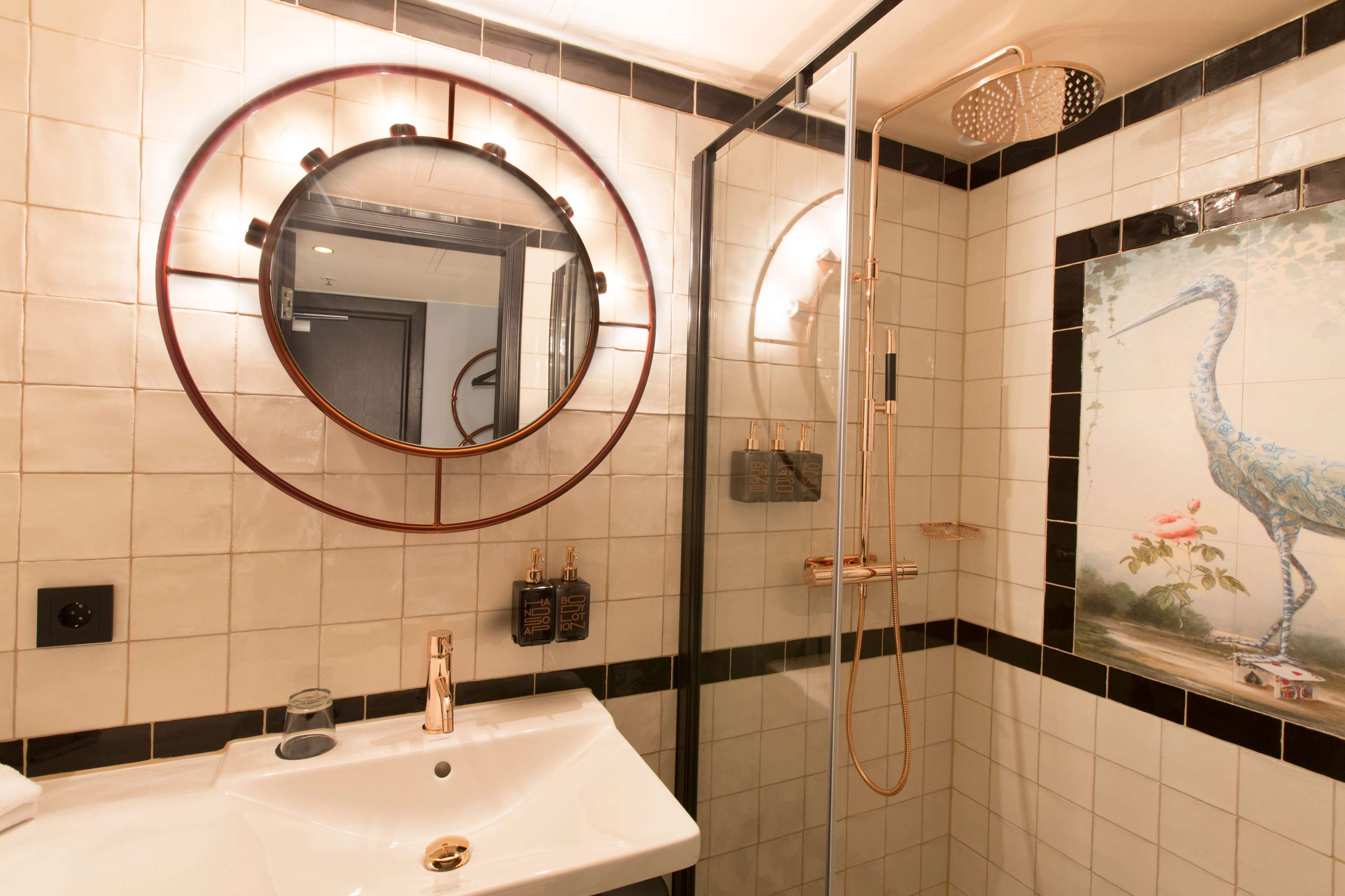 A bathroom with a round mirror, a sink, a shower, and a decorative wall tile featuring a bird.