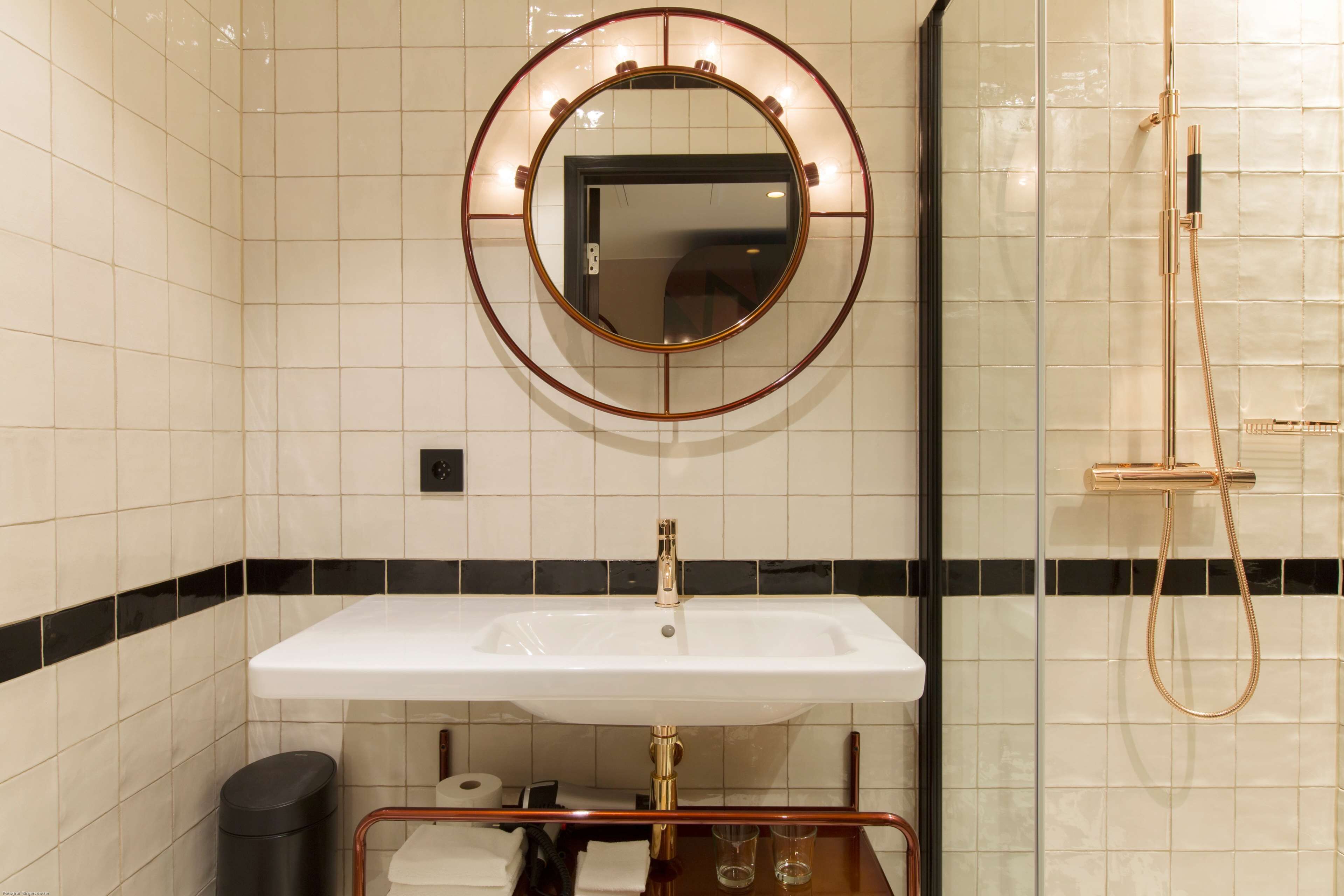 Un baño con un lavamanos blanco, un espejo redondo con iluminación y un cabezal de ducha dorado.