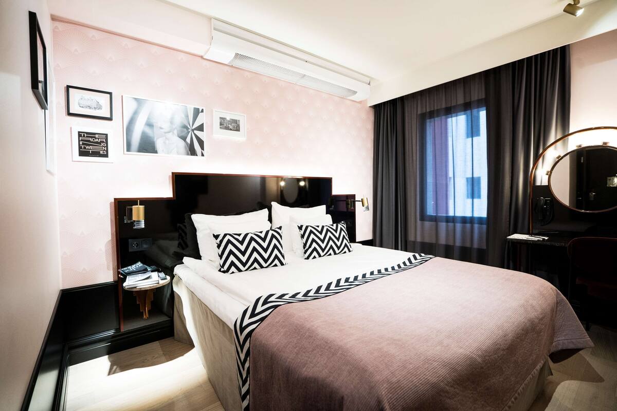 Une chambre d’hôtel moderne avec un grand lit, une table de chevet, un bureau avec une chaise, un miroir et une fenêtre avec des rideaux.