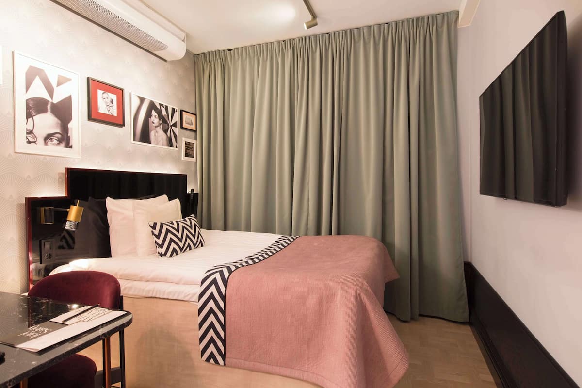 Une chambre d’hôtel avec un lit, un bureau et une chaise. Il y a des rideaux à la fenêtre et un téléviseur fixé au mur.