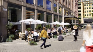 Un café al aire libre con mesas y sillas bajo sombrillas, un edificio con amplios ventanales y un paseo peatonal.
